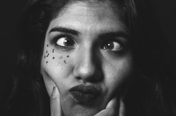 A black-and-white picture of a femme face. They are crossing their eyes, and a hand - possibly their own - is squeezing their cheeks. The right side of their face is covered with small stickers of stars and crescent moons. Image by Jorge Rojas taken under the Unsplash license.